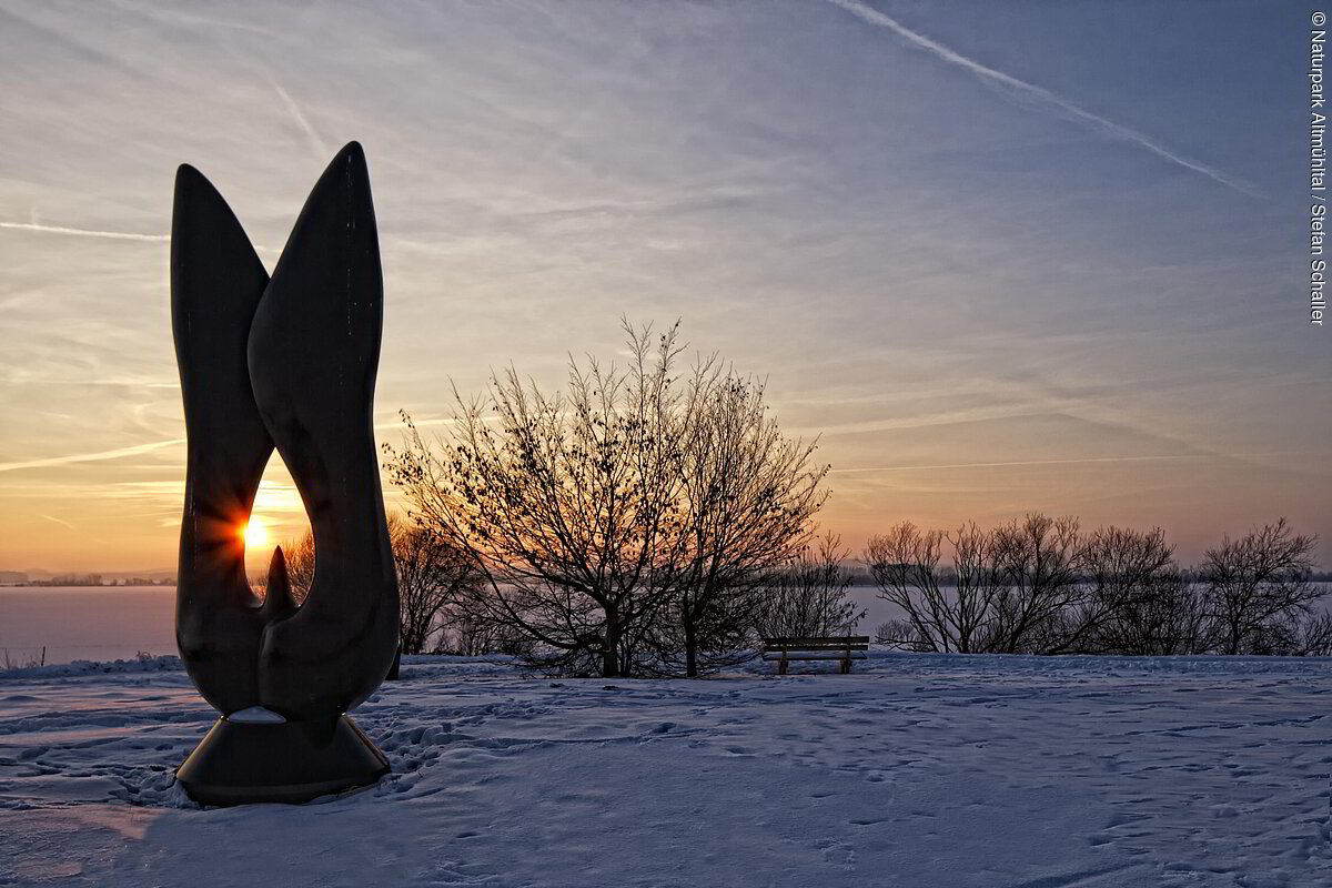Kunstobjekt bei Sonnenaufgang am schneebedecktem Altmühlsee. Einige Meter dahinter stehen einige Sträucher.