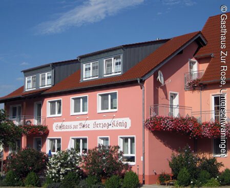 Seitenansicht des dreistöckigen Gasthauses "Zur Rose" in Unterschwaningen mit rötlichen Fassaden und roten Dachziegeln. Im Vordergund blühende Blumen.