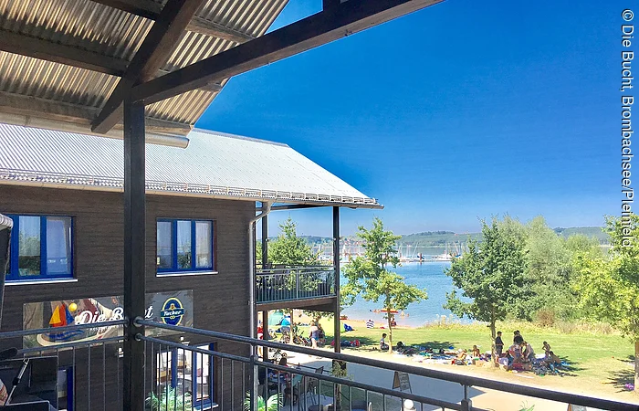 Blick von einem Balkon auf ein zweistöckiges Holzhaus mit Terasse. Dahinter eine Liegewiese mit See.