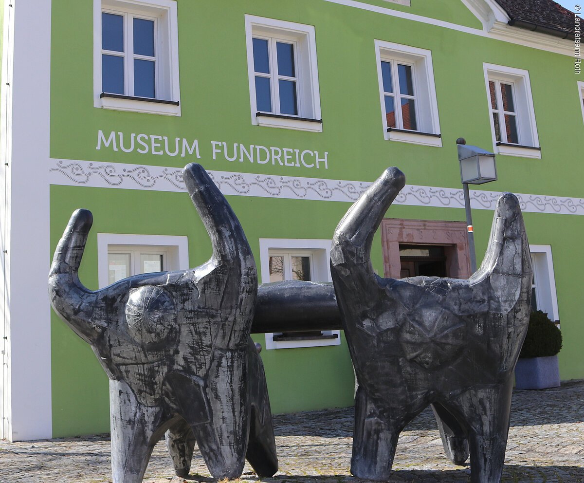 Außenansicht des Archäologischen Museums in Thalmässing. Grünes Haus mit schwarzem Dach. Davor steht eine Skulptur von menschlichen Figuren, die eine Platte tragen.