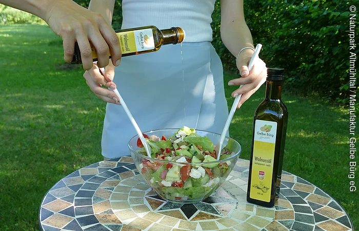 Auf einem runden Mosaiktisch steht eine Glasschüssel mit Salat und eine Flasche Walnussöl. Eine Frau bereitet den Salat zu.