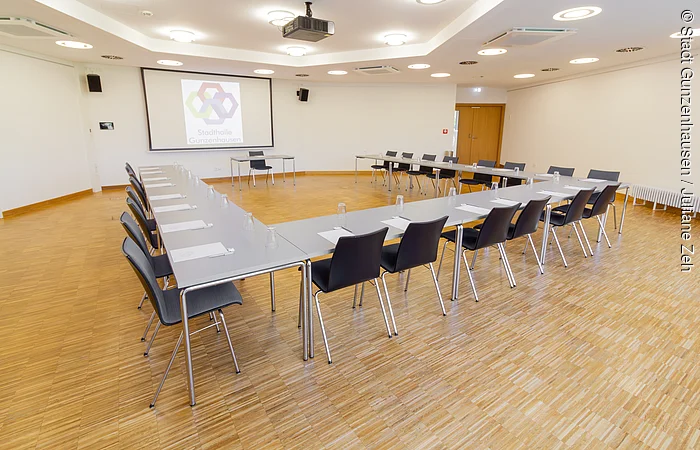 Konferenzraum mit U-förmiger Tischanordnung, Stühlen, Notizblöcken und Gläsern, Projektor und Leinwand an der Wand.