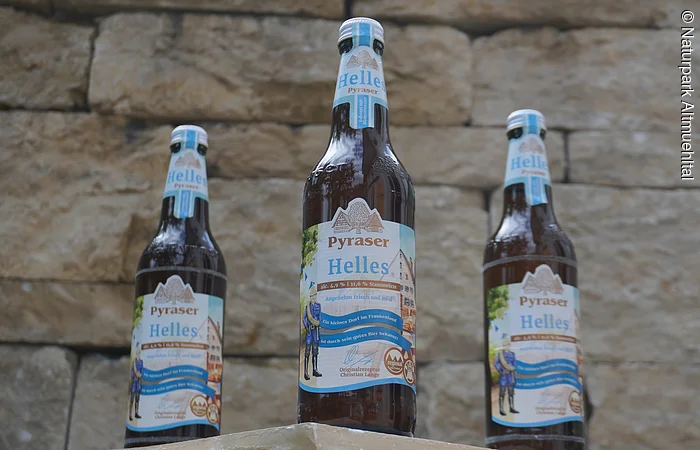 Drei Bierflaschen mit dem Etikett "Pyraser Helles" vor einer Steinmauer.