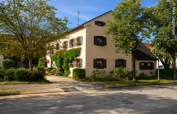 Das Familienhaus ist einen hellen Ton angestrichen und hat grüne Fensterläden, an den Fenstern befinden sich Blumenkästen mit Geranien. Um das Haus befinden sich verschiedene Sträucher und an der westlichen Häuserfront befinden sich Pflanzen