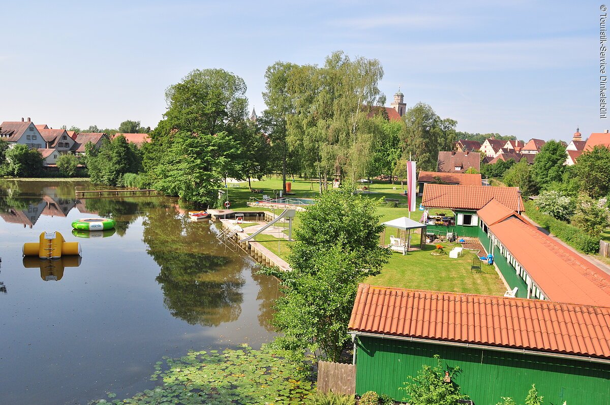 Naturfreibad in Dinkelsbühl Blick auf das Wörnitzstrandbad in Dinkelsbühl. Links das Schwimmbecken des Naturfreibads. Rechts ein grünes Haus entlang des Schwimmbeckens. Im Hintergrund sind Häußer der Stadt Dinkelsbühl.