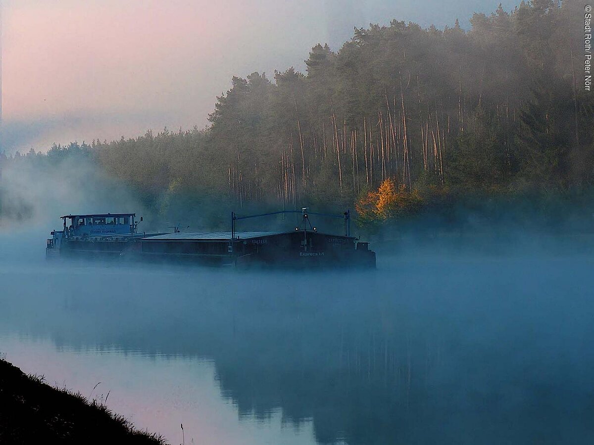 Ein Frachtschiff auf dem Main-Donau-Kanal. Das Wasser leuchtet hellblau. Über dem Kanal hängt Nebel. Am hinteren Ufer erstreckt sich ein Wald der im Nebel liegt.