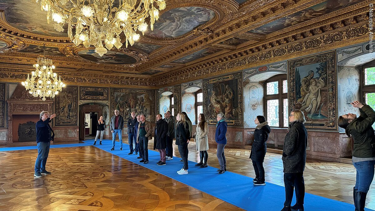 Prunksaal Schloss Ratibor Roth Personengruppe in einem prunkvollen Raum mit verzierten Wänden und Decken, zwei Kronleuchtern und blauen Teppichen.