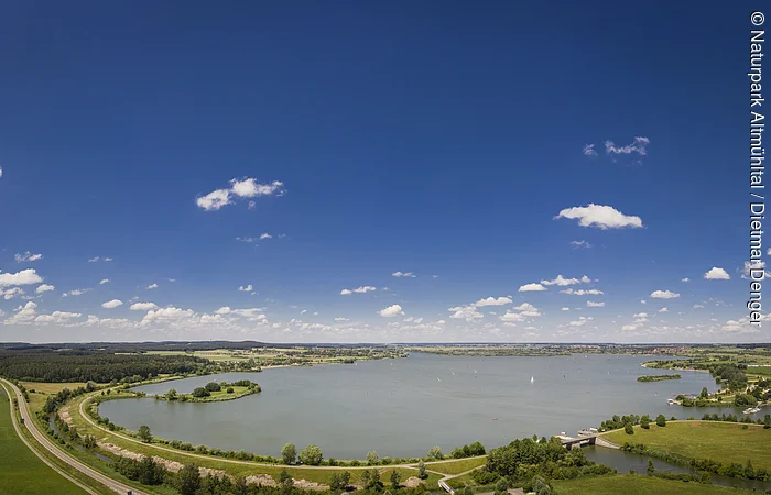 Panoramablick auf einen See mit Uferwegen, Wiesen und blauem Himmel mit vereinzelten Wolken.
