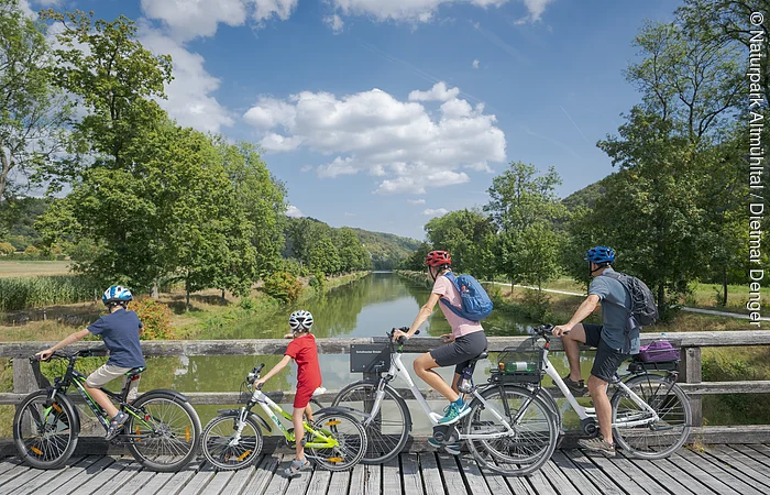 Vier Personen mit Fahrrädern auf einer Holzbrücke mit Blick auf einen Fluss und grüne Landschaft.