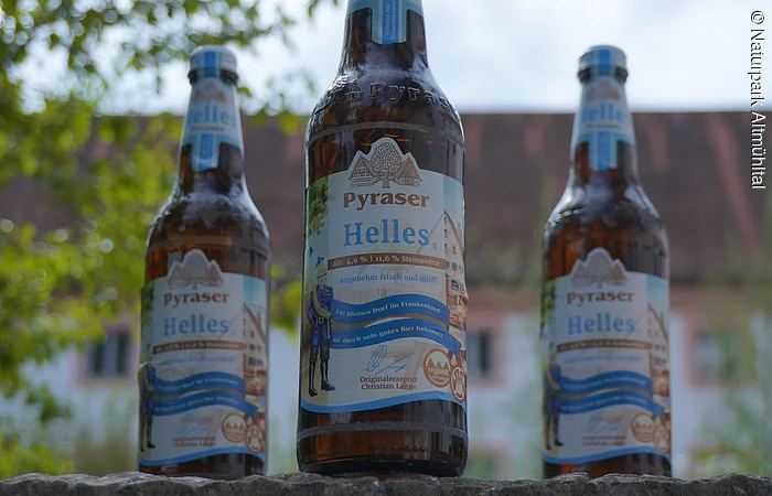 Drei Flaschen Pyraser Helles Bier stehen auf einer Mauer, im Hintergrund sind Bäume und ein Gebäude.
