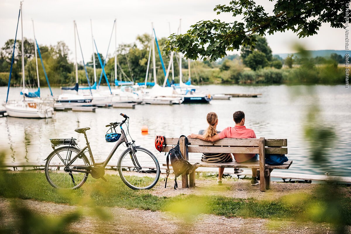 Pärchen am Altmühlsee Altmühltal-Radweg Paar sitzt auf Holzbank am See, Fahrrad mit Helm lehnt daneben, Segelboote im Hintergrund sichtbar.
