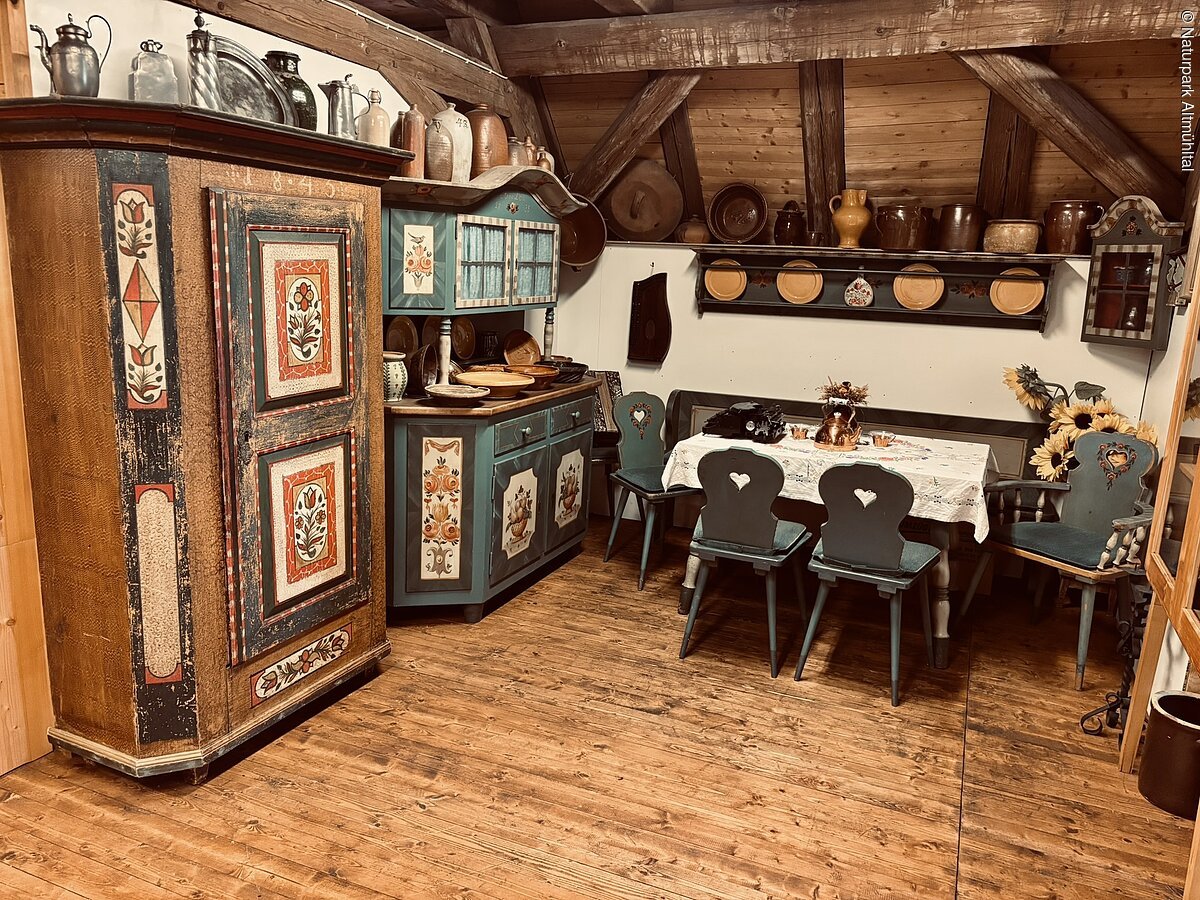 Links sind zwei alte Bauernschränke mit bunten Verzierungen zu sehen. Rechts ein Tisch mit Stühlen