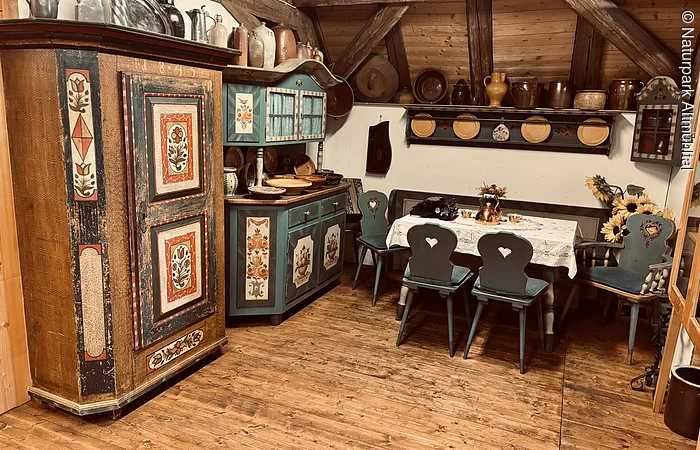 Links sind zwei alte Bauernschränke mit bunten Verzierungen zu sehen. Rechts ein Tisch mit Stühlen