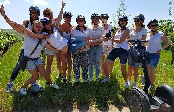 SegwayTouren Eine Gruppe von jungen Frauen mit Helm posiert erfreut für ein Gruppenbild vor einem Weinberg. Vor Ihnen steh ein Segway.