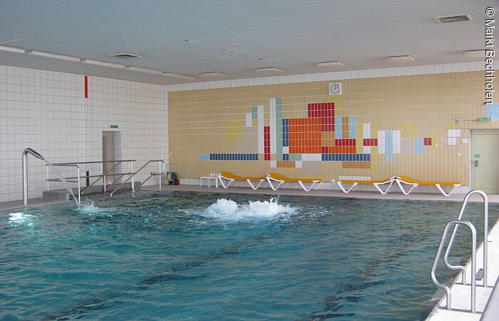 Blick auf die hintere Hälfte eines rechteckigen Schwimmbeckens des Hallenbads in Bechhofen. Das Schwimmbecken nimmt den gesamten Raum ein. Die Wände im Hintergund sind links weiß und rechts bunt gekachelt.