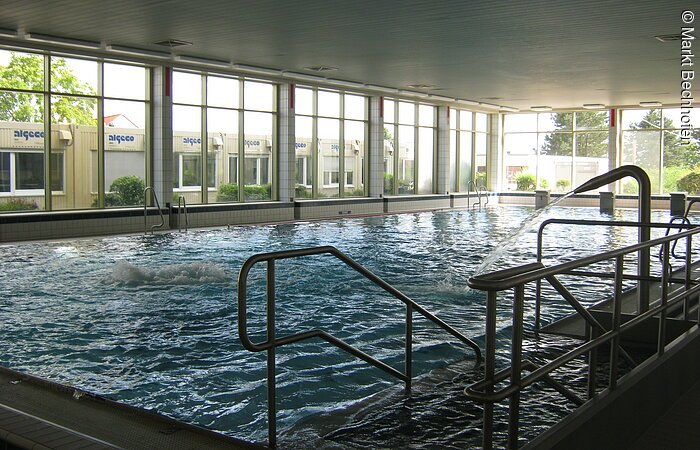 Ein rechteckiges Schwimmbecken des Hallenbads in Bechhofen. Im Vordergrund Geländer und Einstiegstreppe. Im Hintergund erstreckt sich über die gesamte Wand eine Fensterfront.