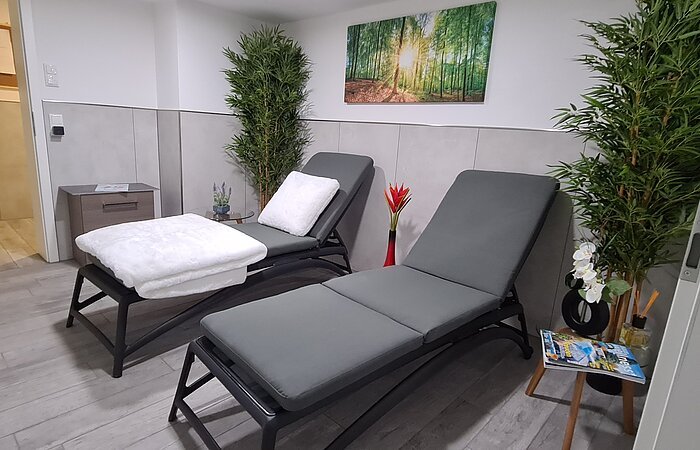 Wellnessbereich Zwei graue Liegen in einem grau geflißeten Bereich. Darüber an der Wand ein Bild mit grünen Bäumen.