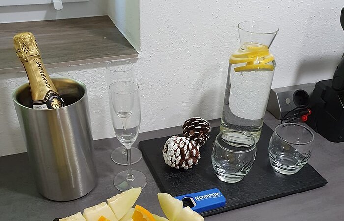 Eine graue Küchenzeiel darauf ein Obstplatte mit schön plaziertem Obst. Dahinter ein Sektkühler mit einer Flasche Sekt, daneben zwei Sektgläser. Rechts davon eine Karaffe Wasser und zwei Gläser.