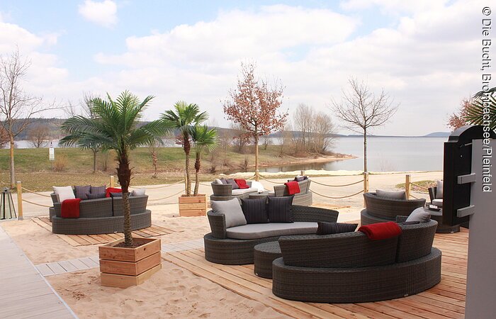 Gemütliche braune Couchen mit Palmen auf einer Holzterasse und Sand. Mit Ausblick auf den Brombachsee.