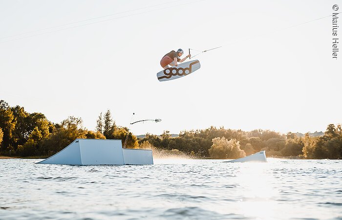 Wakeboardanlage Ein Mann mit Wakeboard springt über ein Hinderniss im Wasser. Dahinter am Uder Bäume und Sträucher.