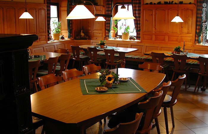 Ein Gastraum mit holzvertäfelten Wänden. In der Mitte des Raumes ein großer Holztisch mit Stühlen. An den Wänden entlang eine lange Bank mit weiteren Tischen und Stühlen.