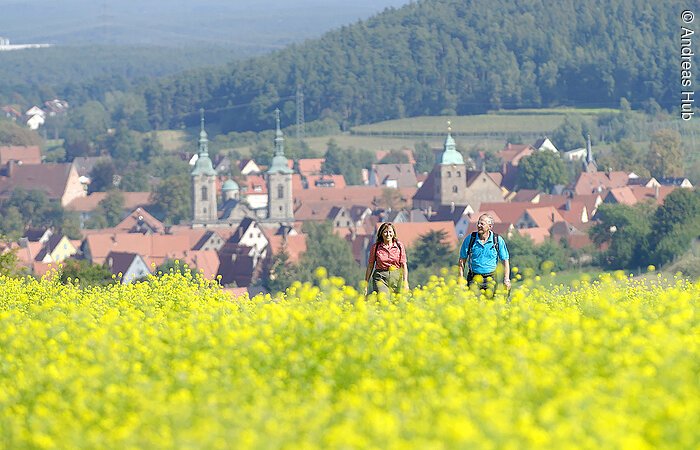 Ein Ehepaar wandert durch ein gelbes Rapsfeld. Im Hintergund die Häuser eines Dorfes.
