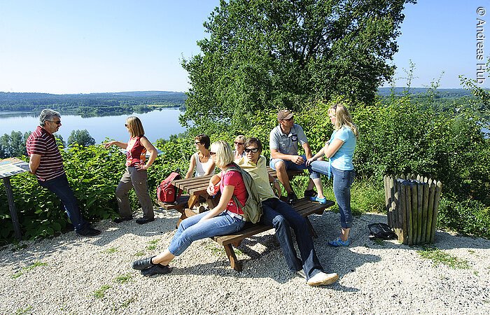Eine Gruppe Wanderer macht Rast auf einer Sitzbank im Grünen. Im Hintergund ein See.