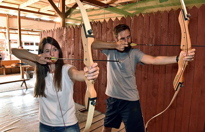 Bogenschießen im Fränkischen Seenland Eine Frau und ein Mann halten jeweils einen gespannten Bogen und zielen mit dem Pfeil. Im HIntergudn ein hoher Holzzaun.