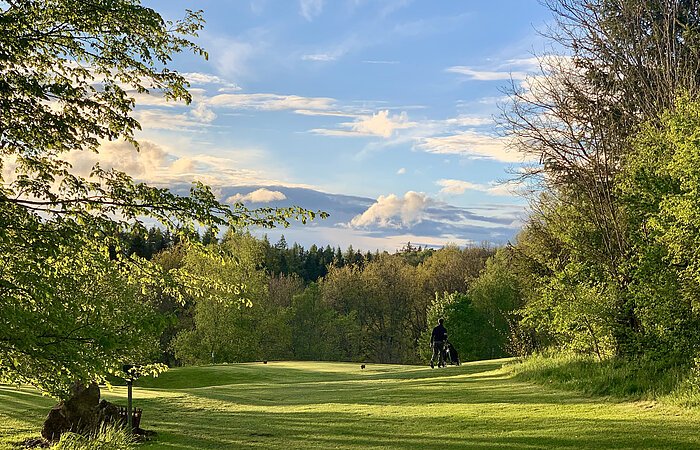 Bahn 10 Blick auf einen Golfplatz. Eine flache grüne Wiese. Links und rechts Bäume und Sträucher. Ein Golfer mit Golftasche läuft über den Platz.