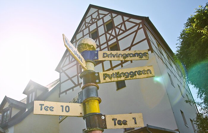 Im Hof der Zollmühle Blick von unten auf einen Schildermast mit goldenen Schildern. Darauf steht in der Reihenfolge von oben nach unten: Drvingrange, Kurzplatz, Puttingarden Tee 10 und Tee 1. Im Hintergund ein großes Fachwerkhaus mit braunen Balken