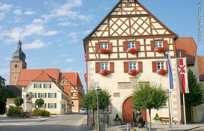Im Fokus ein Rathaus im Fachwerkstil mit roten Balken und gelber Fassade. Vor dem Rathaus ein Krautbrunnen mit zwei Figuren und Krautsköpfen. Im Hintergund ragt der Kirchturm aus den Häusern der Altstadt.