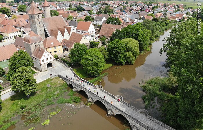 Luftbild der Stadt Ornbau. Eine lange Steinbrücke führt über einen überschwemmten Fluss zu einem Stadttor. Dahinter weitere Häuser einer Altstadt. Kleiner im Hintergund weitere Wohngebiete mit Häusern.