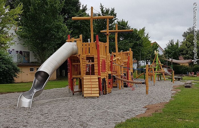 Freizeitpark "Am Bruckespan" Ein Klettergerüst mit Metallrutsche. Das Klettergerüst ist aus Holz. Einzelne Holzbalken sind bunt lackiert. Das Klettergerüst hat die Form eines Segelschiffs.
