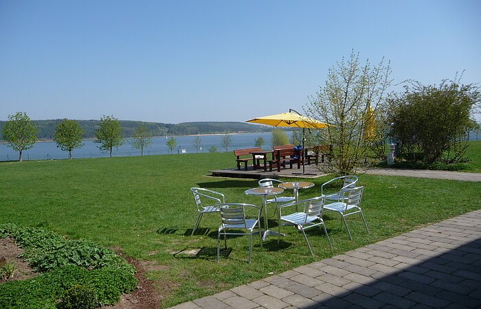 Pause am grünen Ufer Eine Sitzgruppe auf einer grünen Wiese am Ufer eines Sees. Weiter hinten ein Sitzgruppe aus Holz mit gelbem Sonnenschirm.