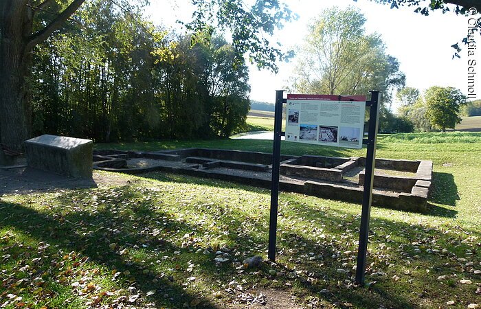 Informationstafel des Panorama-Rundwanderwegs steht auf einer grünen Wiese. Dahinter eine alte Ruine aus Stein.