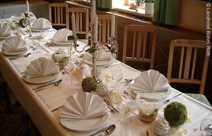Stillvoll gedeckter Tisch mit weißer Tischdecke und gefalteten Servietten. Ein Kerzenleuchter und weiß grüner Dekoration auf dem Tisch. Holzstühle am Tisch.