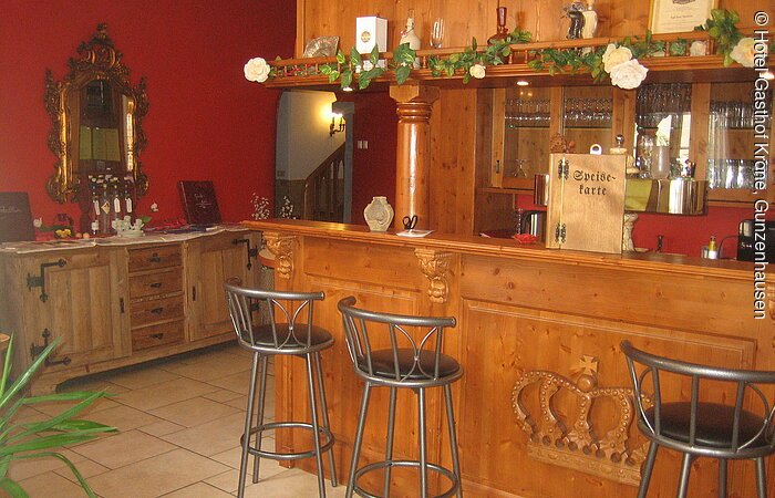 Gasthaus "Zur Krone" Eine Bar aus Holz mit drei Barhockern. In den Regalen hinter der Bar stehen Gläser. Links von der Bar im Hintergund ein Sideboard aus hellem Holz vor einer rot gestrichenen Wand.