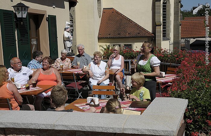 Die Gäste des Gasthof Zur Krone sitzen auf der gut gefüllten Steinterassse. Die Terasse ist geschmückt mit roten Blumen. Im Hintegrund ein kleiner Ausschnitt einer Kirche.