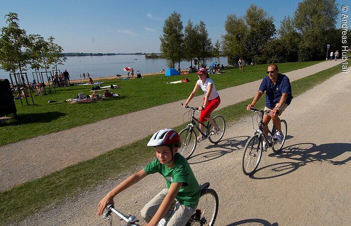 Drei Radfahrer radeln auf einem Fahrradweg am See. Im Hintergund der See mit Sandstrand umgeben von Bäumen.