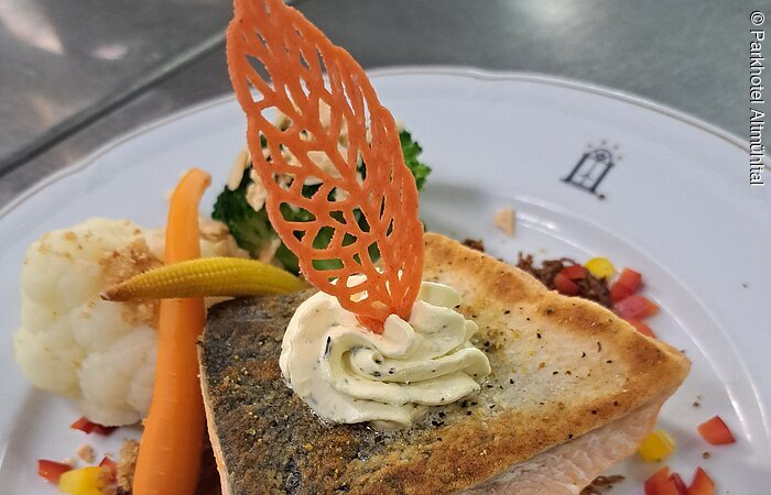 Lachs vom Parkhotel Altmühltal Ein rosa gebratenes Stück Lacks mit einem Blatt und Merrettich dekoriert, liegt auf einem Kartoffelsrösti. Das weiße Teller ist zudem mit Blumenkohl, Karotte und Paprika dekoriert.
