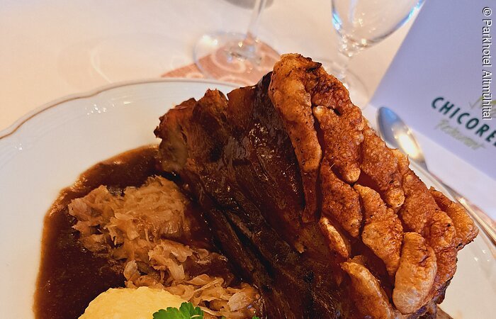 Schäufele vom Parkhotel Altmühltal Zwei Kartoffelknödel mit Petersilie garniert liegen auf Bratensoße. Dahinter ein schönes, knuspriges Stück Schäuferle. Ein Sektglas ist zur Hälfte zu sehen.