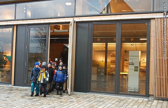Eine Gruppe von Kindern steht am barrierefreien Eingang der Umweltstation des Landesbundes für Vogelschutz in Muhr am See Kinder kommen aus dem Ausstellungsgebäude maschiert.
