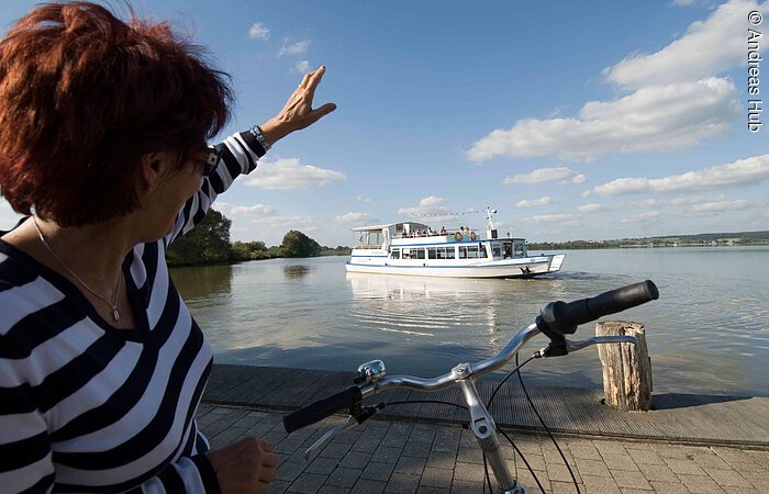 Eine Fahrradfahrerin winkt vom Ufer aus der MS Altmühlsee zu.