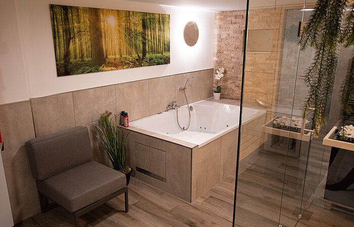 Ein modernes Badezimmer mit großer Badewanne. Daneben ein grauer Sesel. An der Wand hängt ein Bild mit Bäumen. Im rechten Bildrand eine Glastür einer Dusche.