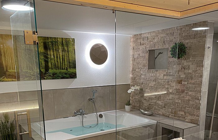 Blick aus einer Sauna durch eine Glastür auf eine große Badewanne. Ein beige gefließter Wellnessbereich. An der Wand ein grünes Bild mit Bäumen.