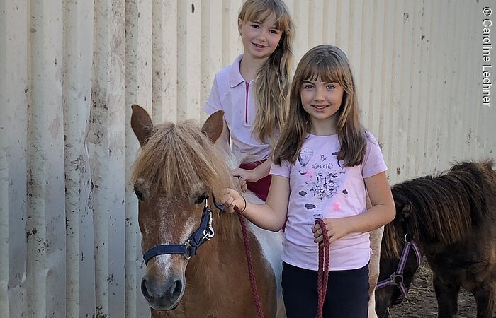 Zwei junge Mädchen in rosa Reitkleidung mit zwei Ponys vor einer blechernen Stallwand. Ein Mädchen sitzt auf einem Pony. Das andere steht daneben. Beide Mädchen lächeln in die Kamera.