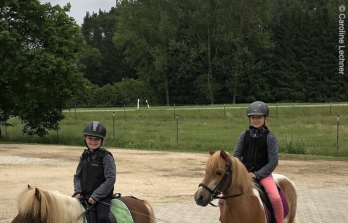 Zwei junge Mädchen sitzen in voller Reitkleidung auf zwei kleinen Ponies. Die Tiere stehen auf einem gepflasterten Hof. Beide Mädchen lächeln in die Kamera. Im Hintergund ein grüne Weide.