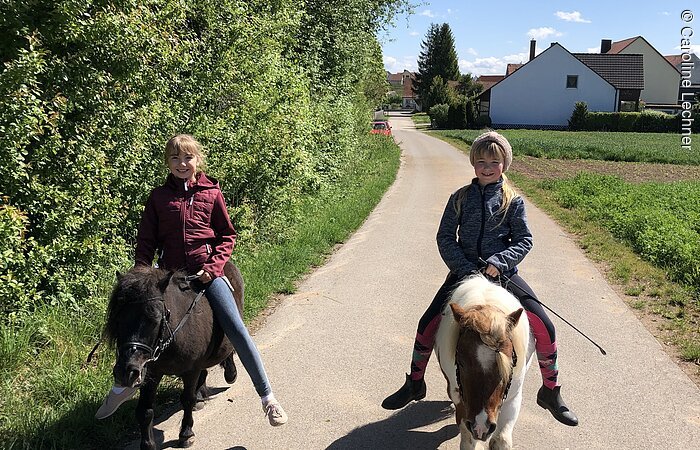 Zwei junge Mädchen reiten auf zwei kleinen Ponies auf einem geteerten Weg entlang einer Böschung. Beide Mädchen lächeln in die Kamera. Im Hintergund ein Wohnhaus.