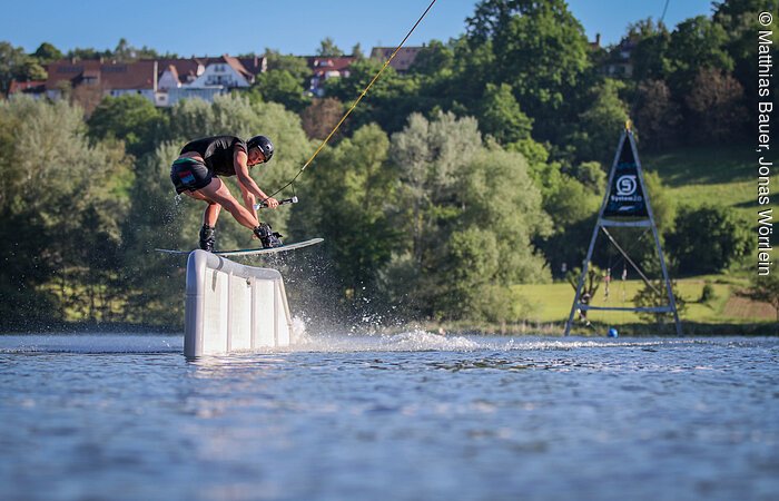 Eine Wakeboarderin fährt mit ihmer Wakeboard über ein Hindernis im See. Am hinteren Ufer grüne Bäume und Sträucher.