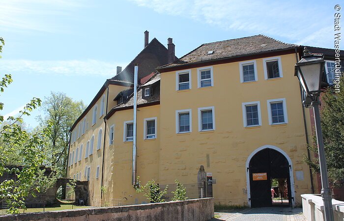 Schloss im Wörnitzgrund Außenansicht eines Schlosses mit gelber Fassade. Ein hohe Gebäude mit mindesten vier Ebenen. Den Eingang bildet ein großes Holztor. Eine Steinbrücke führt zum Eingang.